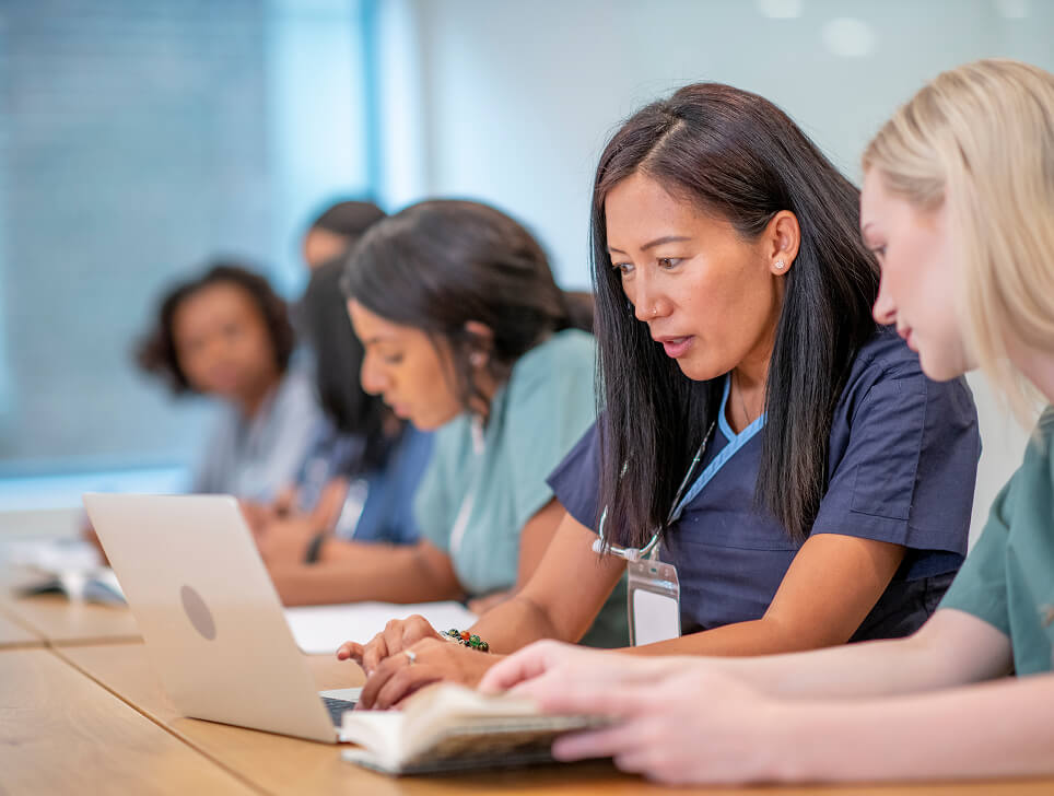 A diverse group of women in scrubs, look over a laptop and notes at a conference table.