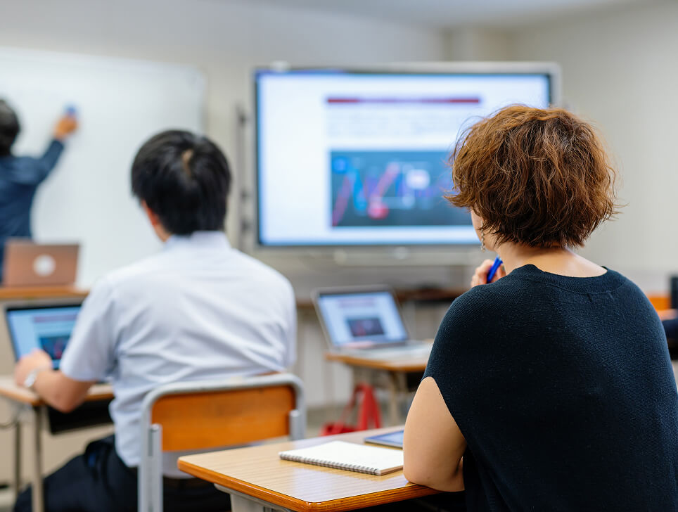 People in a classroom observing someone writing on a whiteboard next to a screen.