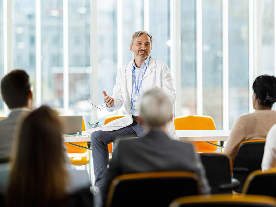 A doctor in a white coat sits on a desk, addressing a small, seated audience.